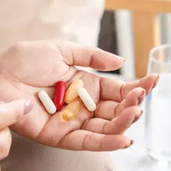 Dłoń z kilkoma kapsułkami, w tym czerwona i żółta. Obok szklanka wody i butelki z lekami. Kiedy probiotyki szkodzą, warto zastanowić się nad ich przyjmowaniem.
